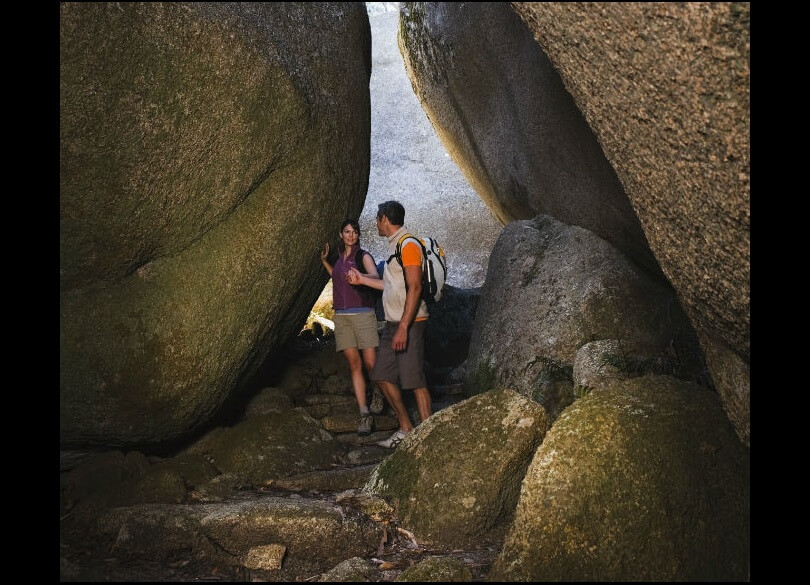 Two bushwalkers standing on a rocky path between 2 large boulders.