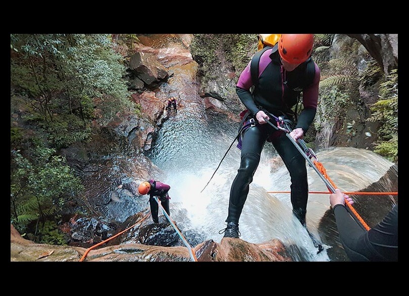 Two people abseiling down a waterfall, one has just started to descend, and the other is almost at the base.