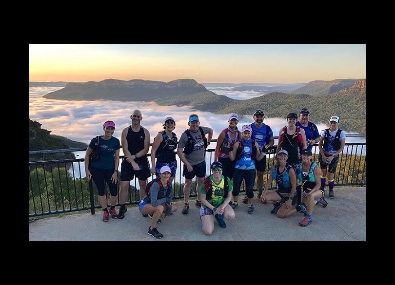 A group of runners standing at a scenic overlook. In the background, the majestic Blue Mountains rise, and below them, a layer of clouds stretches out.