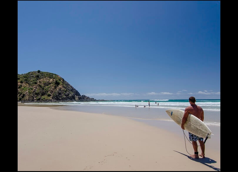 A person holding a surfboard looks out towards the ocean on a sunny beach day, with clear blue skies above, waves gently crashing near the shore, and a rocky hill to the left. 