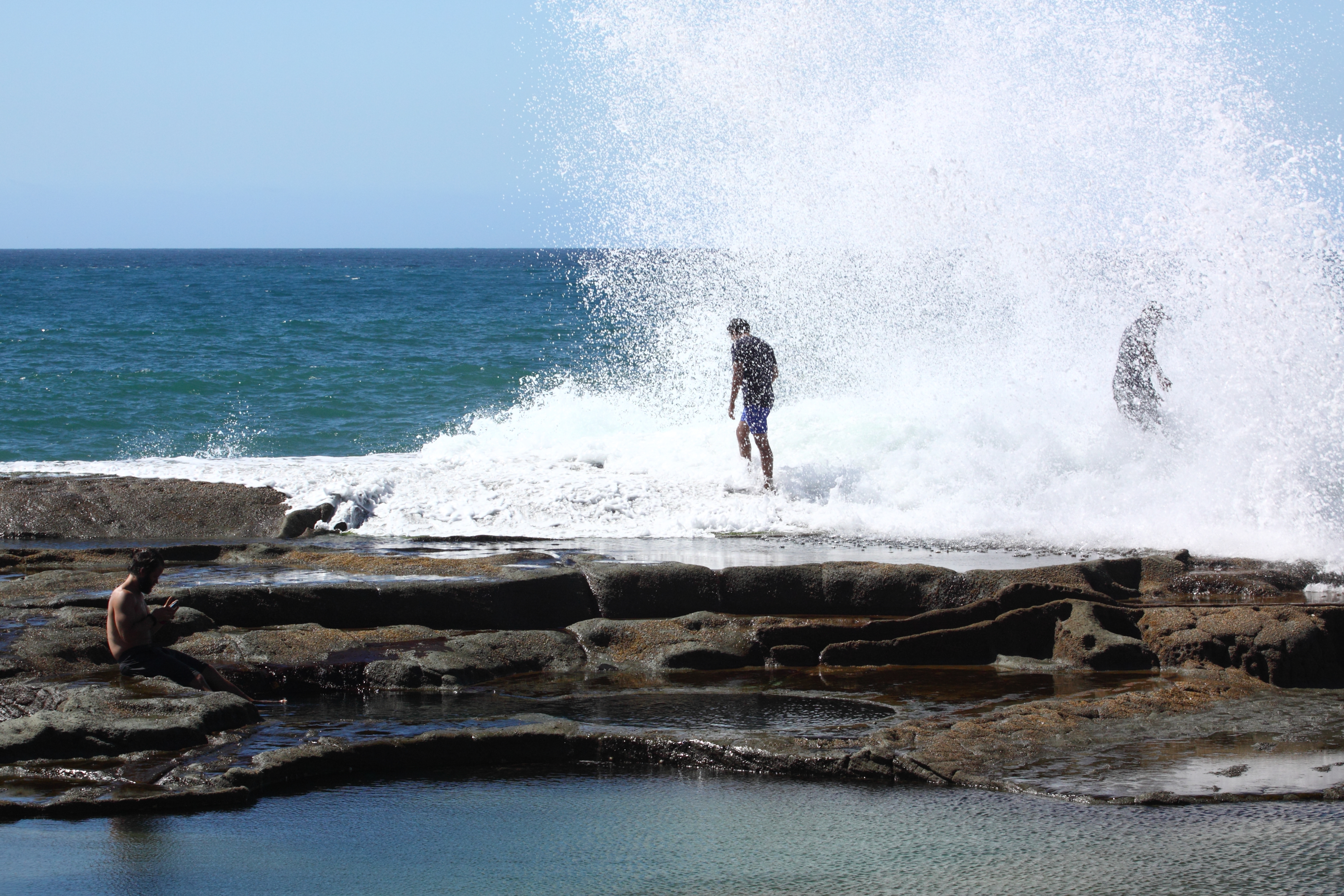 Two men are standing on rocks by the ocean and a large wave has crashed against the rocks covering them in water.