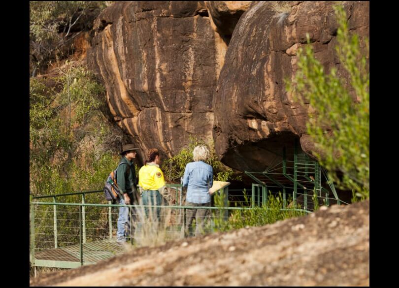 Three individuals on a metal viewing platform examining a stratified brown rock formation surrounded by green vegetation.