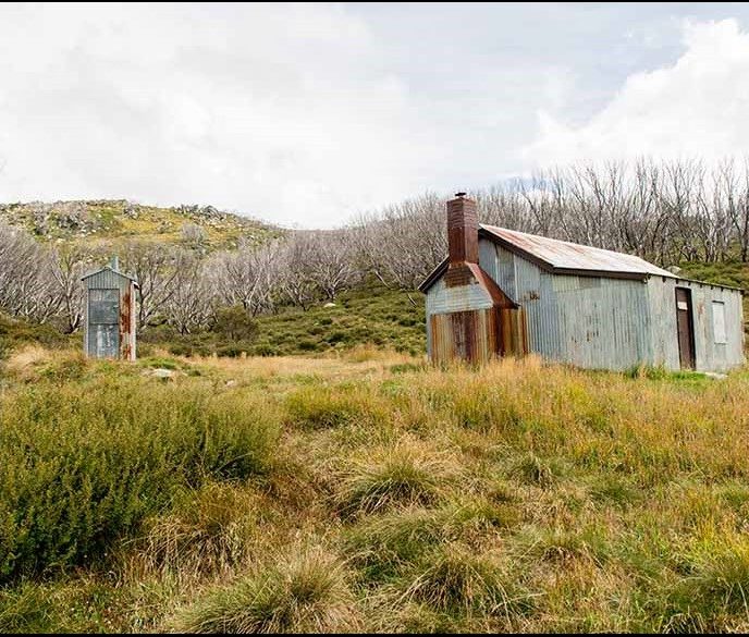 Schlink Hut, a rustic metal-clad mountain shelter with a chimney, set in the grassy landscape of Kosciuszko National Park with sparse trees and rolling hills in the background.