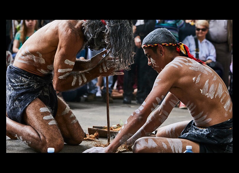 Two Aboriginal men crouched down while starting a fire. They are painted in traditional custom while a crowd of people watch.