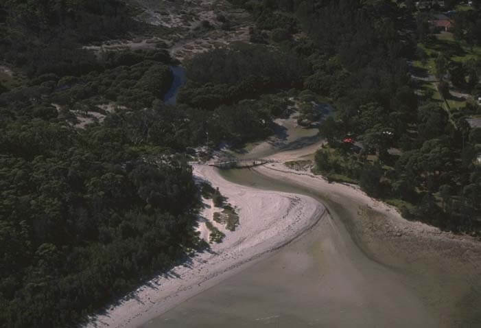 A view of Callala Creek meeting the sea surrounded by tree-lined sand dunes and Callala Beach in the foreground