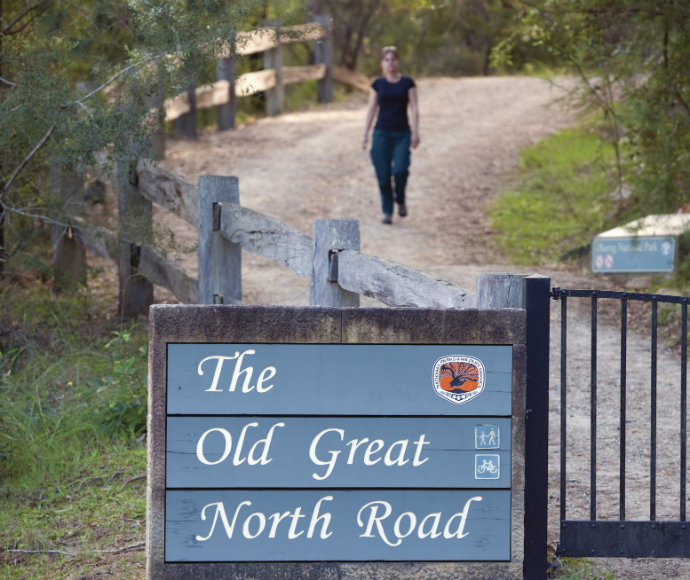 A female tourist walks along the road behind the sign for the Old Great North Road in Dharug National Park.