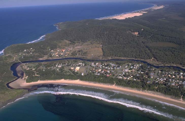 Aerial view of Korogoro Creek winding through a dense green landscape and flowing into the ocean. The creek is bordered by a curving sandy beach, with a small community of buildings near the water’s edge and a long stretch of beach backed by sand dunes and vegetation on the opposite side.