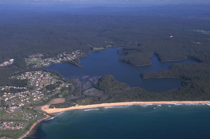 This stunning aerial view of Lake Mummuga highlights the calm blue waters of the lake, beautifully framed by lush green forests. Nearby, you can see a welcoming coastal town and a sandy beach that gently meets the ocean. It's a wonderful example of nature and community coming together