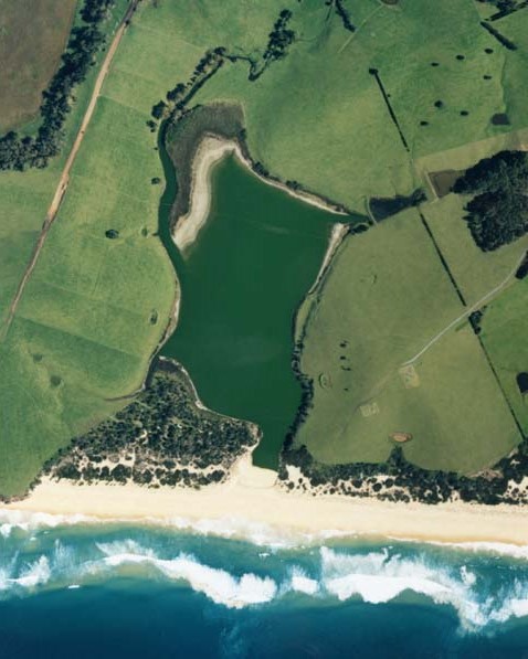 Aerial view of Little Lake, Wallaga, showing a vibrant green lake adjacent to a sandy beach with waves crashing onto the shore. The lake is surrounded by lush fields with varying shades of green and patches of trees. The contrast between the calm lake waters, the dynamic ocean waves, and the structured agricultural land provides a diverse and captivating landscape.