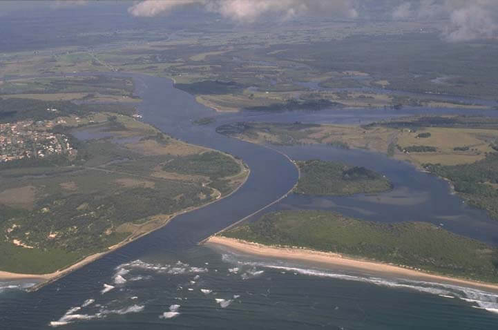 An aerial view of a wide river winding through a landscape of green land areas with patches of development, including buildings and clearings. The river flows into the sea, with a distinct line where the darker river water meets the lighter blue ocean. A sandy beach separates part of the river from the ocean on one side. The sky is partly cloudy, casting shadows on the land and water below.