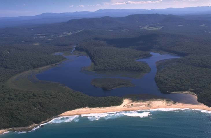 Aerial view of Meroo Lake Estuary showing a winding body of water surrounded by dense greenery, with a beachfront meeting the ocean at the bottom edge.