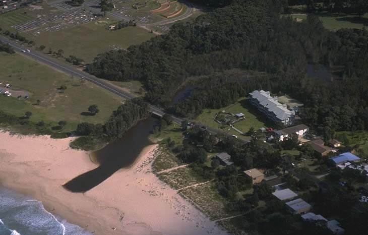 Aerial view of Mollymook Creek flowing into the ocean, with a sandy beach on one side and lush greenery surrounding the water. A bridge crosses over the creek, and buildings are nestled among trees near the water’s edge.