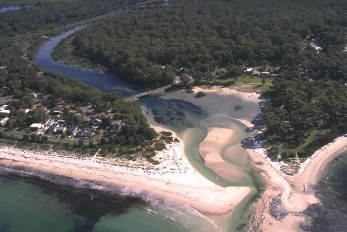 Aerial view of Moona Moona Creek, showing its winding path as it flows into the ocean. The creek is surrounded by a dense residential area with numerous houses and streets, and a beach running parallel to the coastline. The contrast between the natural waterway and developed land highlights the intersection of human habitation and natural landscapes.