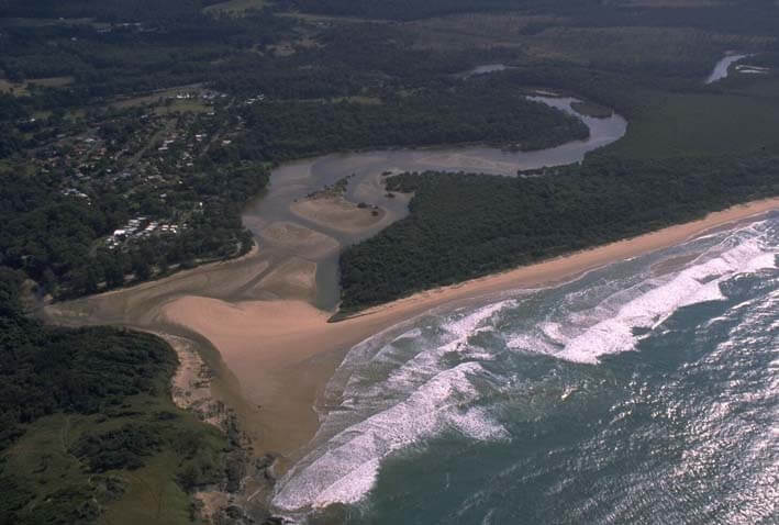 Aerial view of Moonee Creek, showing its winding path as it flows into the ocean. The creek is surrounded by a dense residential area with numerous houses and streets, and a beach running parallel to the coastline. The contrast between the natural waterway and developed land highlights the intersection of human habitation and natural landscapes.