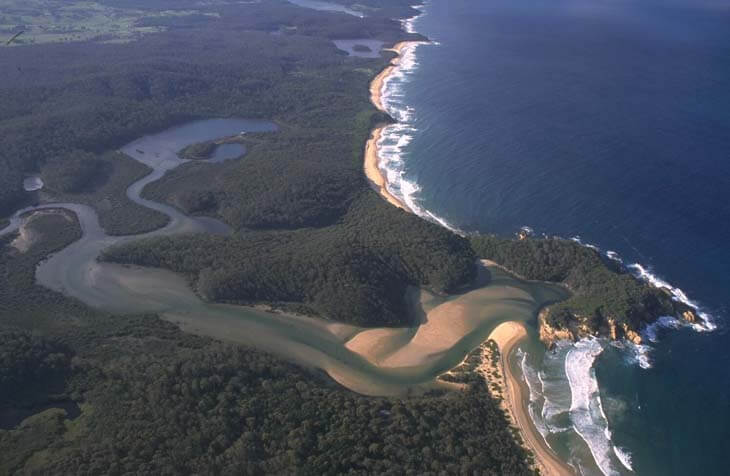 Aerial view of Nelson Lagoon flowing into the ocean, with visible sandbanks at the river mouth and a nearby town along the waterways.