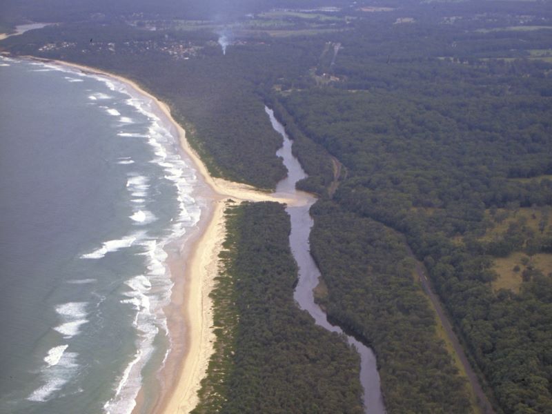 Aerial view of Oyster Creek, featuring a winding river flowing through dense greenery and emptying into the ocean. The creek meanders from the top left corner to the bottom center of the image, where it meets a beach that stretches along the right side. The surrounding landscape is a mix of dark green forest areas and lighter green open spaces. The water appears dark, contrasting with the white surf along the shoreline.