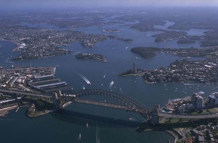 Aerial view of Sydney Harbour, showcasing the iconic Sydney Harbour Bridge spanning across the center. The bridge connects the central business district on the right to a residential area on the left. Multiple boats are visible on the water, indicating a bustling maritime hub. The vast blue waters are dotted with numerous small land masses, and urban development encroaches upon green spaces, illustrating the coexistence of human engineering marvels and natural beauty.