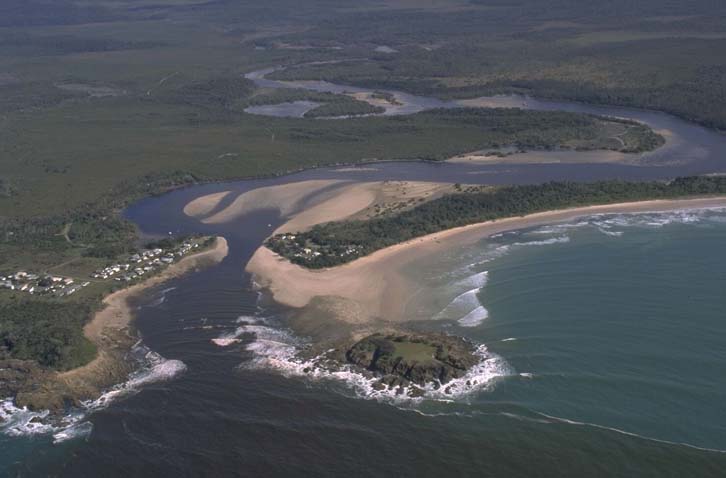 Aerial view of Sandon River winding through a green landscape before meeting the sea. The river’s wide mouth features visible sandbanks, contrasting with the dark blue ocean. Dense greenery and a small settlement are near the river’s mouth.