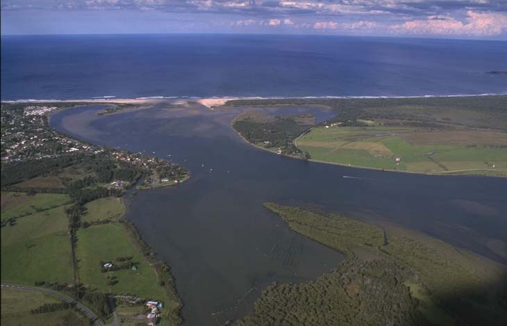 Aerial view of Shoalhaven River winding through a green landscape before meeting the sea. The river’s wide mouth features visible sandbanks, contrasting with the dark blue ocean. Dense greenery and a small settlement are near the river’s mouth.