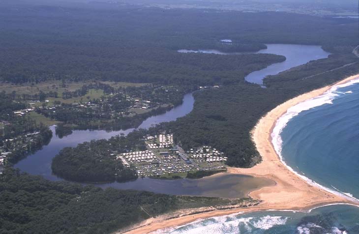 Aerial view of Tabourie Lake showing a winding water body with varying shades of blue, surrounded by dense green vegetation. To the right, a sandy beach borders the lake, leading into the open sea with waves visible along the shoreline. A cluster of buildings is nestled between the lake and the beach, indicating a small coastal community. The contrast between the calm lake waters, structured human settlement, and dynamic ocean waves highlights the diverse natural and human-influenced landscapes.