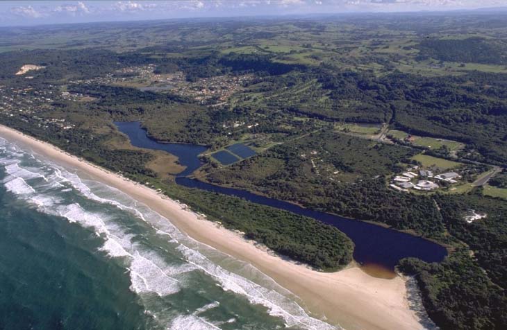 Aerial view of Tallow Creek near a sandy beach with waves from the ocean on one side, surrounded by green vegetation and nearby buildings indicating human habitation.