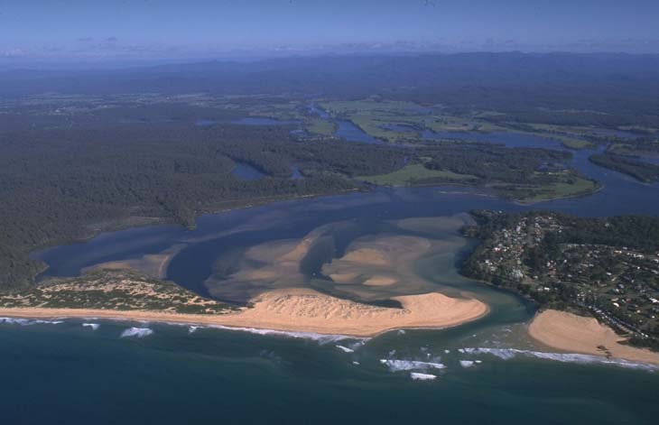 Aerial view of the Turros River winding its way to the ocean. The river’s brownish water meets the blue sea, with sandy beaches forming interesting patterns at the mouth. Surrounding the river are patches of greenery and some developed areas with buildings and roads near the coastline.