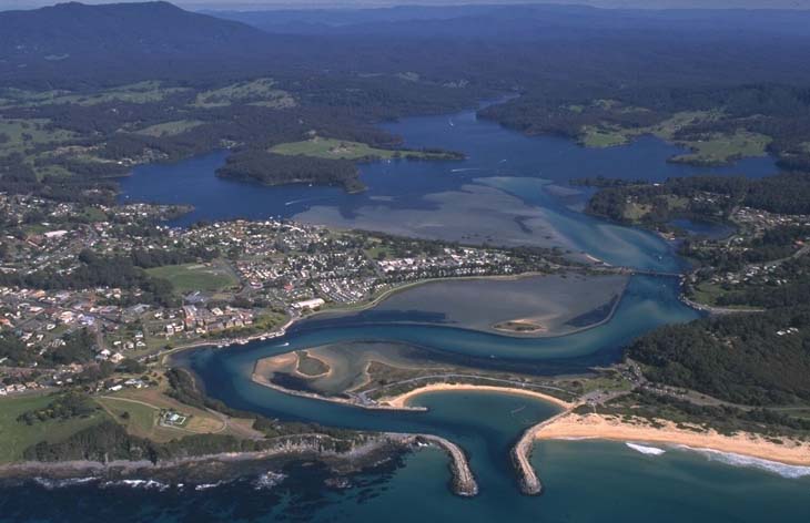 Aerial view of Wagonga Inlet showcasing a coastal town with a mix of residential and commercial buildings, streets, and a marina. The town is surrounded by dense greenery and forested areas. The bay itself is a large body of water with a few boats visible, and the coastline features sandy beaches and rocky outcrops. In the distance, there are rolling hills and mountains under a partly cloudy sky, adding depth to the landscape.