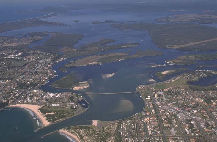 An aerial view of Wallis Lake estuary, featuring a large body of water with multiple branching inlets and outlets. The estuary is surrounded by a mix of dense urban development and natural green landscapes. The water is calm, with several visible sandbanks. The image highlights the interface between urban areas and natural waterways.