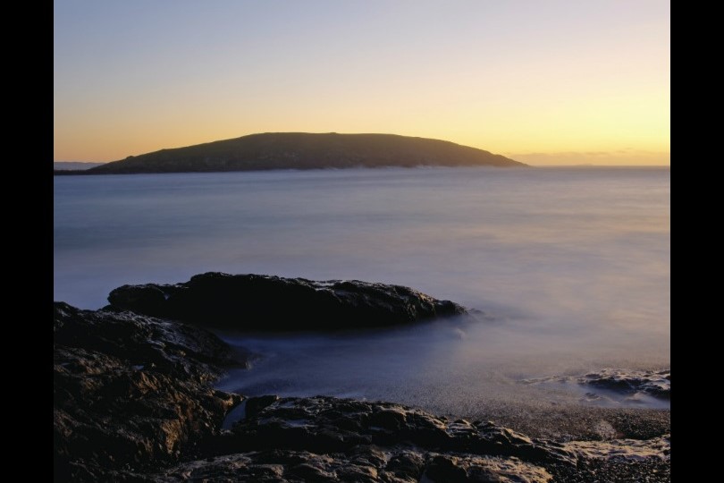 Serene view of Muttonbird Island Nature Reserve at dusk, featuring rocky shores with gentle waves and the island’s silhouette against a gradient sky transitioning from warm yellow to cool blue tones.