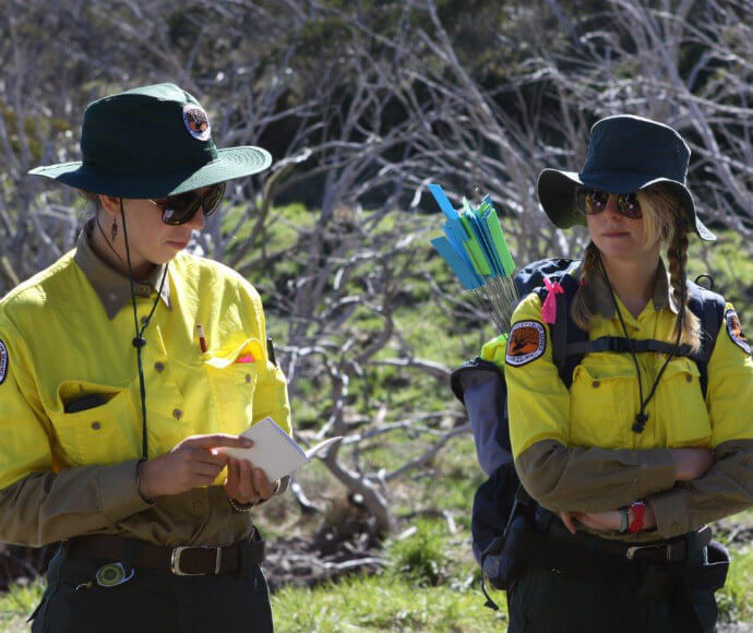 Two Park Rangers in uniform; one is writing on a notepad and the other carries arrows in their backpack, standing in a natural outdoor setting.