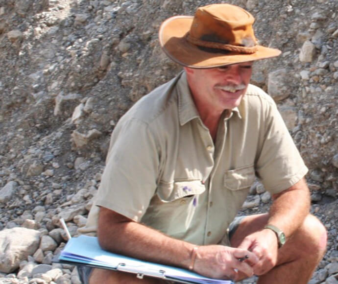 A man wearing a brown hat and khaki top, crouched down looking at soil