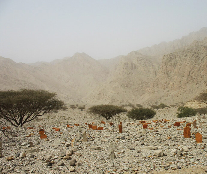 a landscape of grey rocks with grey mountains in the background, but there are red rocks stood upright marking graves