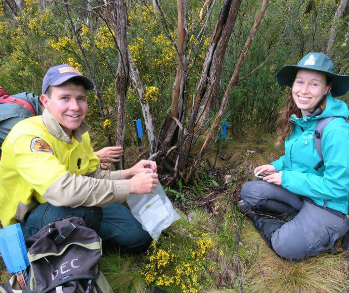 Two individuals in outdoor gear and hats are sitting on the ground in a wooded area with shrubbery, one holding a clear plastic bag and the other holding an object.