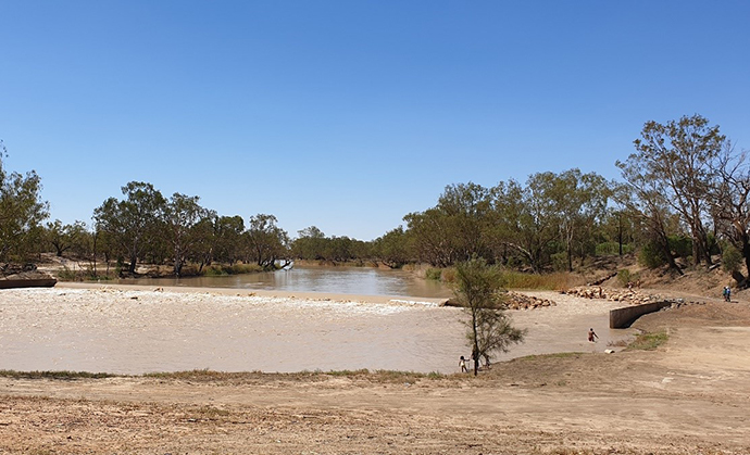 A wide river with a moderate flow of water, flanked by trees and a clear blue sky overhead. A concrete structure is visible on the right bank, and there are people standing near the water’s edge on both sides.