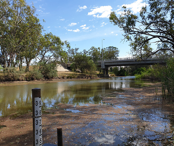 Natural outdoor scene featuring a body of water, possibly a river or lake, with greenery on the banks. In the foreground, there is a measurement gauge indicating depth in meters, ranging from 3.6 to 4.0 meters. A bridge spans across the water in the background under a partly cloudy sky during daytime.