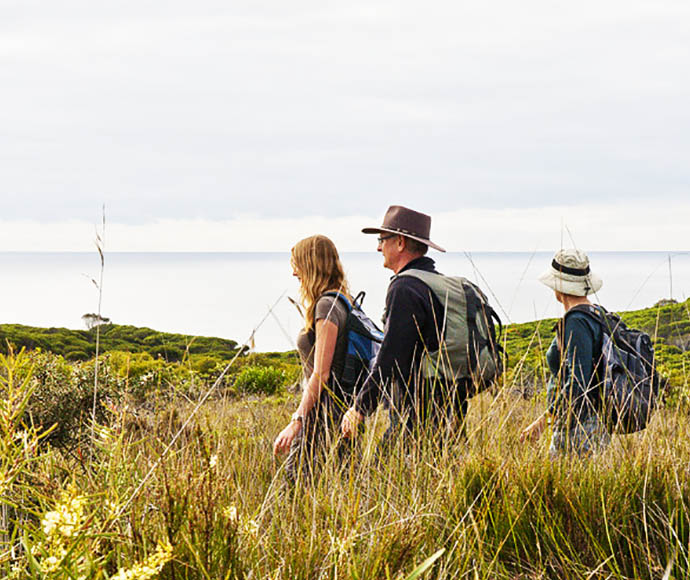 Three hikers with backpacks walking through a grassy landscape with the ocean in the background, on the Light to Light walk in Ben Boyd National Park.