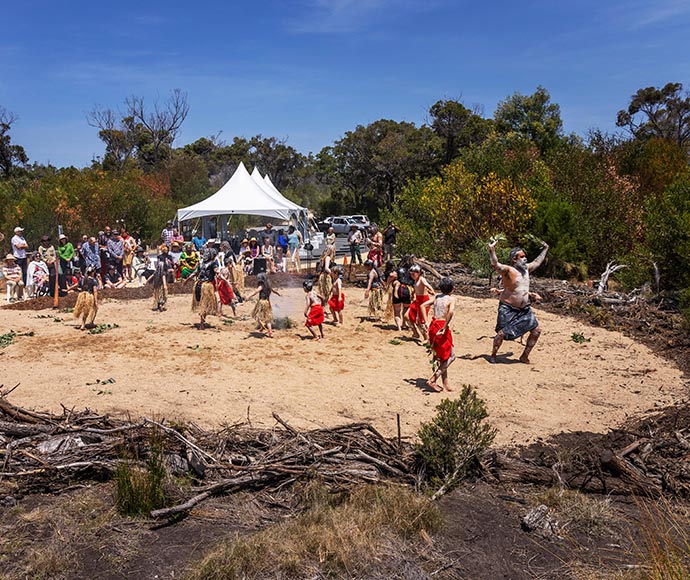 A group of people are gathered in a sandy area, participating in a cultural or traditional dance. There is a white canopy tent in the background, and the surroundings feature green vegetation and trees under a clear blue sky. 