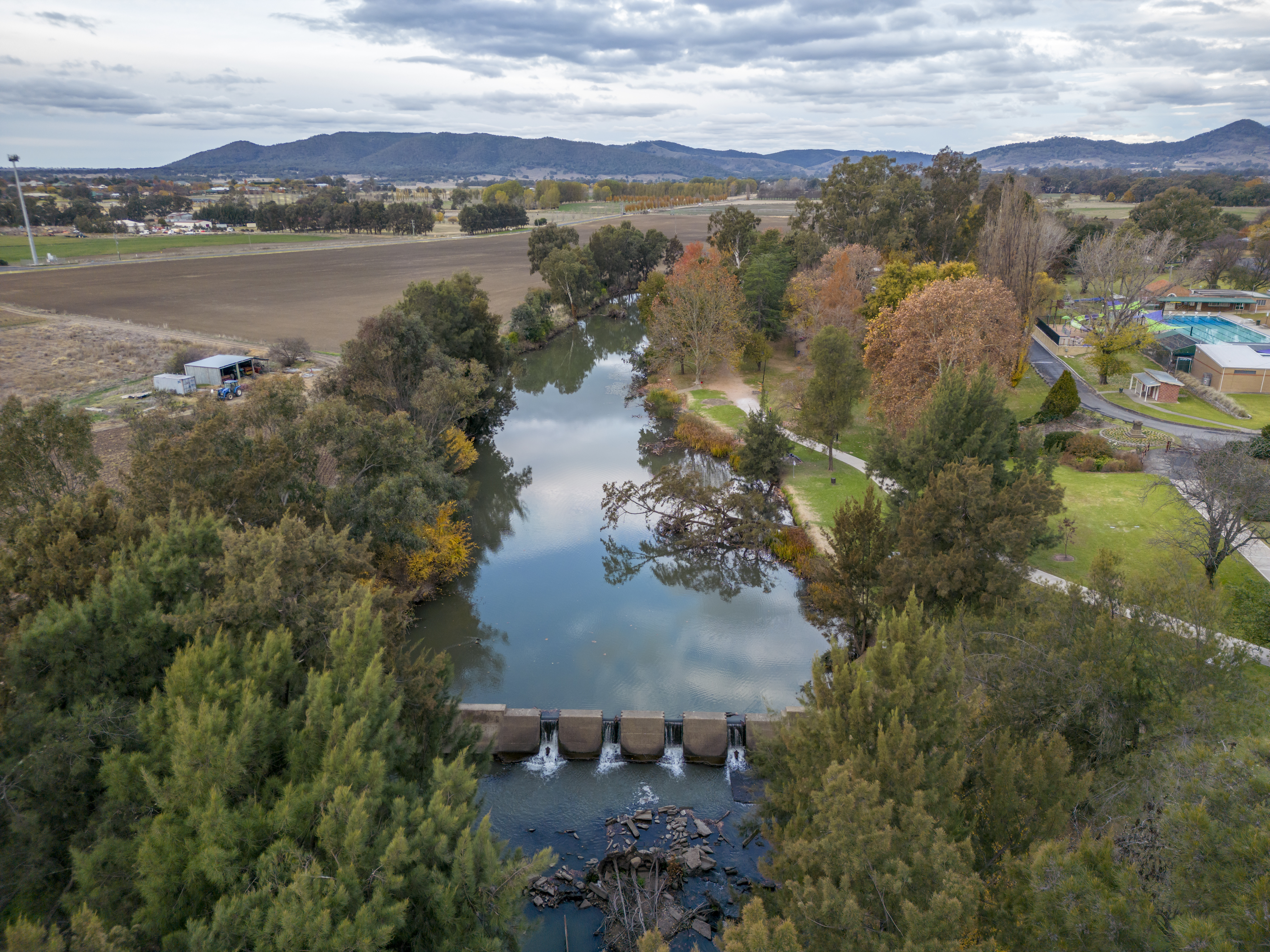 An aerial photo of the Cudgegong River near Lawson Park in Mudgee. The image features a blue, cloudy sky, with abundant vegetation on both sides of the river. A dam is situated in the middle of the river, and mountains can be seen in the background.