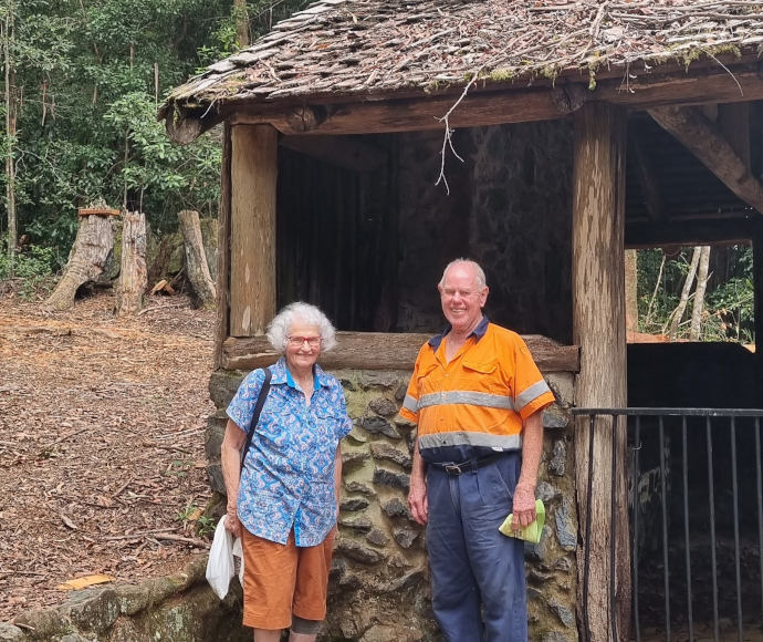 An elderly couple stand in front of a small stone hut with a thatched roof and log frame in a clearing in a forest