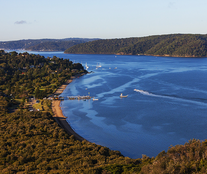 An aerial view of the Barrenjoey Lighthouse Track at Ku-ring-gai Chase National Park. The image showcases the winding track leading to the lighthouse, surrounded by lush greenery and coastal landscapes. The bright blue sky complements the vibrant scenery, highlighting the natural beauty of the area.