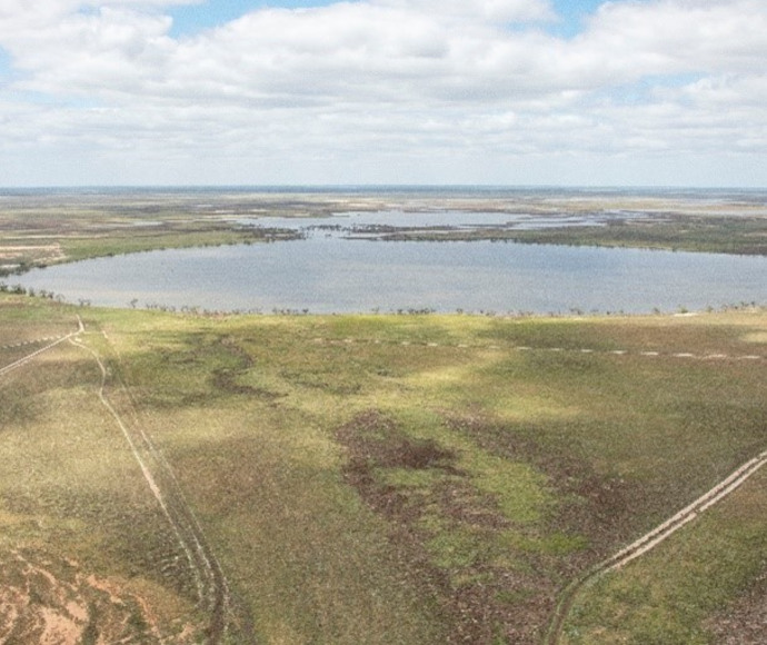 An aerial view of a landscape featuring a large body of water surrounded by greenery and fields with various shades of brown and green, indicating a natural, rural setting.