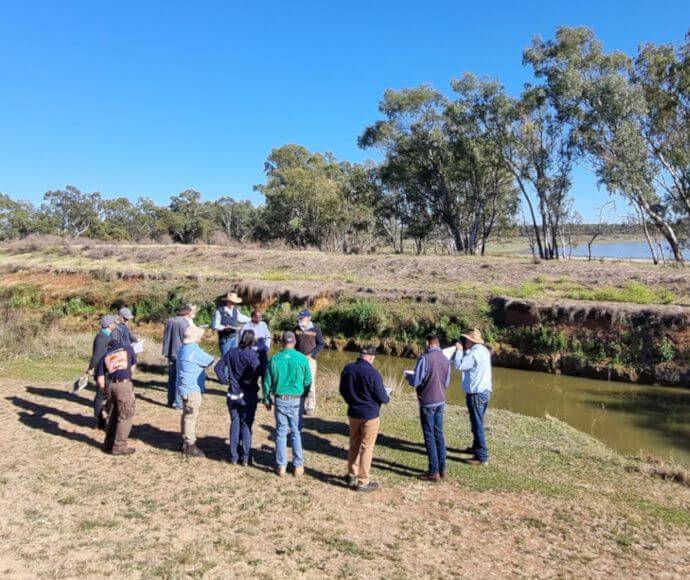 A group of individuals standing on a grassy area near a body of water, possibly engaged in an outdoor educational activity or field trip. The setting includes clear skies and trees in the background, indicating a natural environment.