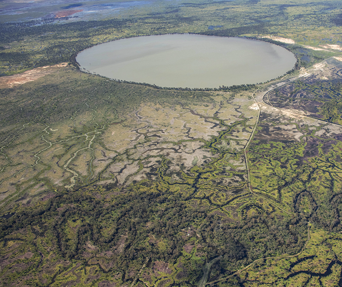 Aerial view of a large circular lake surrounded by green vegetation with intricate patterns of water channels and land weaving through it, indicating a wetland ecosystem. A road runs parallel to the lake on one side, showcasing the contrast between natural and human-made landscapes.