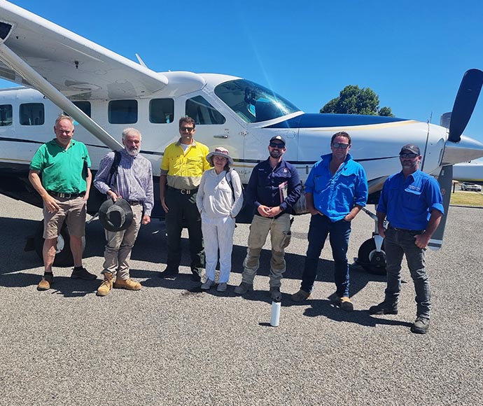 A group of seven individuals standing in front of a white propeller aircraft with blue and black detailing. The individuals appear to be pilots or aviation staff, indicated by their uniforms and headgear. The setting is outdoors on a clear, sunny day.