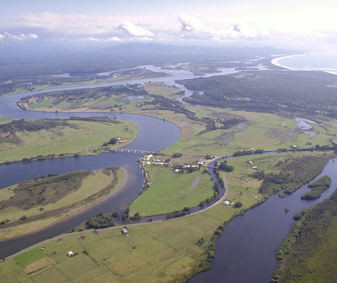 An aerial view of a large river system the land changes colour from green grass to dark green, brown and sandy in the areas where there are mangroves and saltmarsh