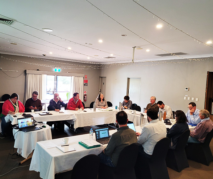 A group of individuals seated around a large conference table in a well-lit room with white walls and carpeted flooring. Some are focused on laptops, while others are listening or reading materials. The room has natural light coming through the windows and festive string lighting along the ceiling’s edge.