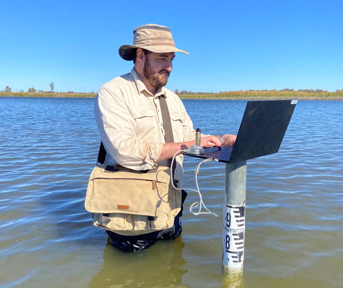 An individual stands in a body of water while using a laptop, with a measuring stick indicating the water depth. The person is wearing a wide-brimmed hat and carrying a shoulder bag, possibly for work-related items. This image highlights an unconventional work setting, possibly for remote work or environmental research.