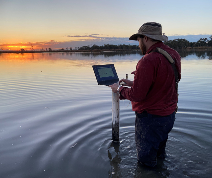 An individual standing in a body of water at sunset or sunrise, holding an open laptop in one hand and a cylindrical object in the other. The sky is painted with hues of orange, yellow, and blue, reflecting on the water’s surface.