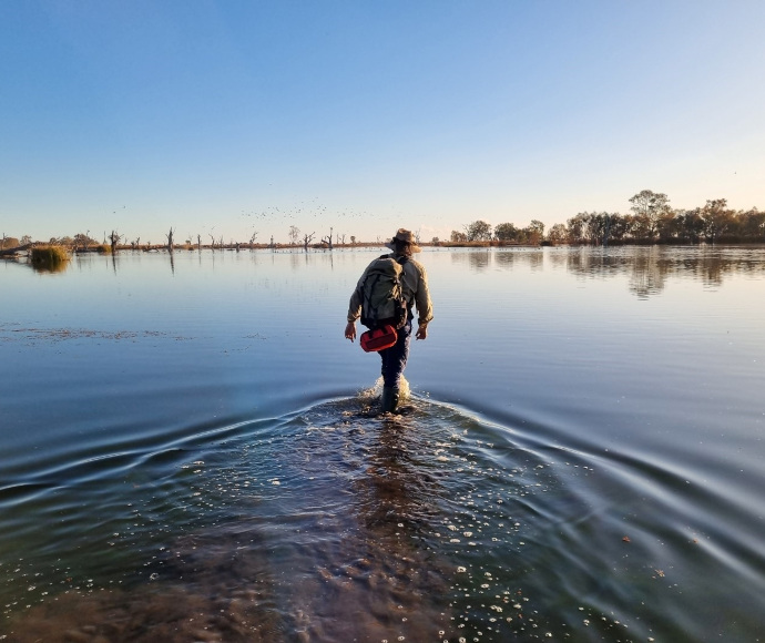 A person standing in calm water up to their ankles, facing away from the camera, wearing a backpack and holding a red object. The horizon shows sparse vegetation and partially submerged trees under a clear sky.