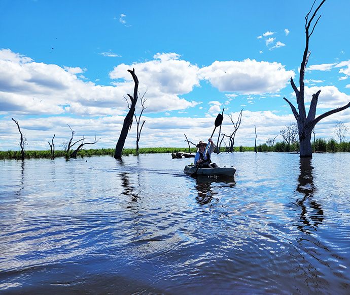 Two individuals kayaking in a body of water with dead trees protruding from the surface, under a partly cloudy sky.